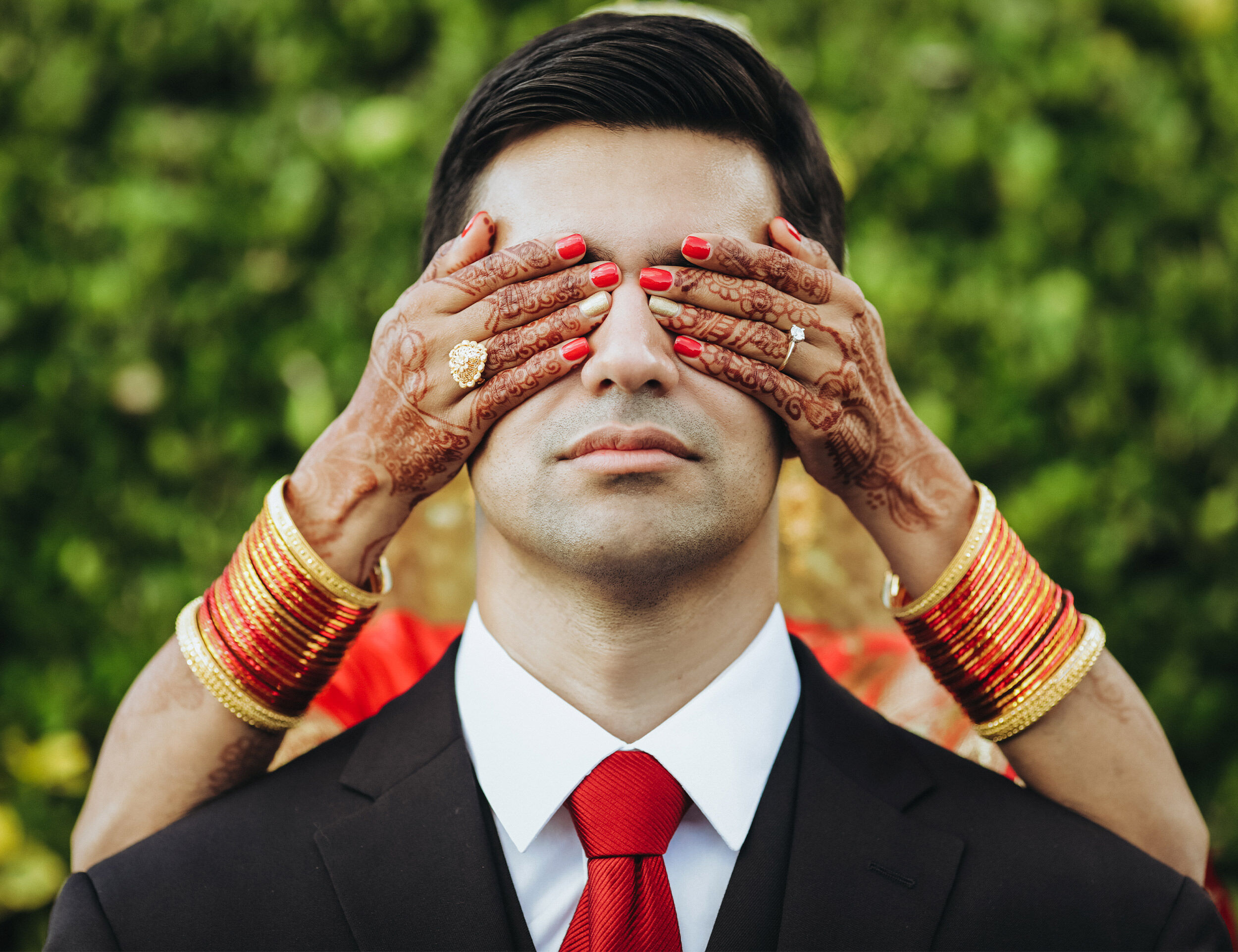 Bride playfully covering groom's eyes with her mehndi-adorned hands during a pre-wedding photoshoot, wearing red bangles and rings